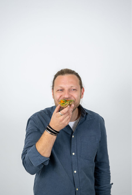 Mann mit langen Haaren im Zopf, der mit einem Lächeln in ein bunt belegtes Vollkornbrot beißt.