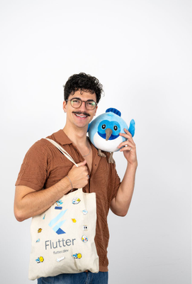 Junger Mann mit Brille und Schnurrbart hält eine bedruckte Stofftasche mit Flutter-Logo in der einen Hand und eine große, blaue Vogel-Plüschfigur an die Wange in der anderen Hand, vor neutralem Hintergrund.