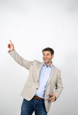 Lächelnder Mann in hellem Sakko und Jeans zeigt mit ausgestrecktem Arm nach oben links.