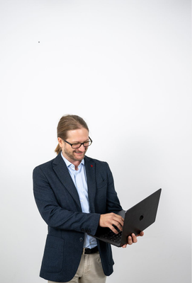 Mann mit Brille und dunkelblauem Sakko tippt auf einem aufgeklappten Laptop und blickt lächelnd auf den Bildschirm.