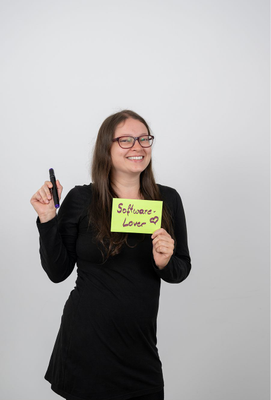 Junge Frau mit langen braunen Haaren und Brille, die lächelnd ein Schild mit der Aufschrift „Software-Lover“ und einem gezeichneten Herz hält; in der anderen Hand hält sie einen schwarzen Marker.