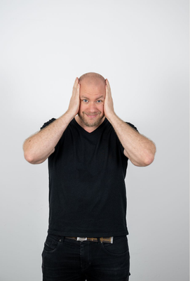 Mann mittleren Alters in schwarzem T-Shirt hält sich mit beiden Händen den Kopf und blickt lächelnd in die Kamera, neutrale Pose vor hellem Hintergrund.