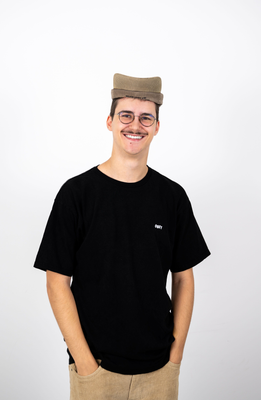 Junger Mann mit rundem Brillenrahmen und Schnurrbart lächelt in die Kamera, trägt ein schwarzes T-Shirt mit „OBEY“-Logo, beiger Kordhose und eine auf ungewöhnliche Weise gefaltete Mütze.