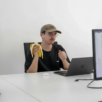 Junger Mann mit Baseballkappe und kabellosen Kopfhörern sitzt vor einem Laptop, hält eine geschälte Banane in der Hand und wirkt konzentriert.
