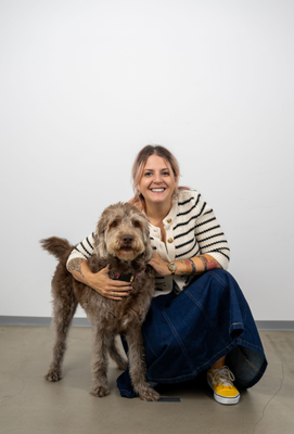 Lächelnde Frau mit rosafarbenen Haaren kniet neben einem zotteligen braunen Hund und legt ihren Arm um ihn; beide blicken in die Kamera.